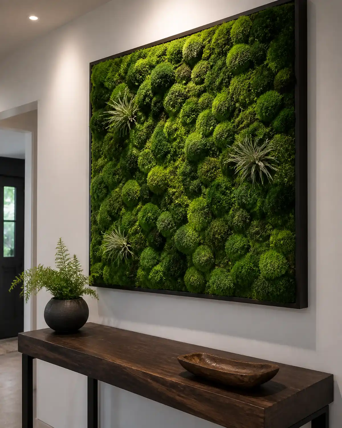 A preserved moss wall art installation in an entryway showing a large framed panel of rich green preserved reindeer moss mounted on a white wall above a console table with a few small air plants tucked into the moss creating striking natural artwork with soft ambient lighting