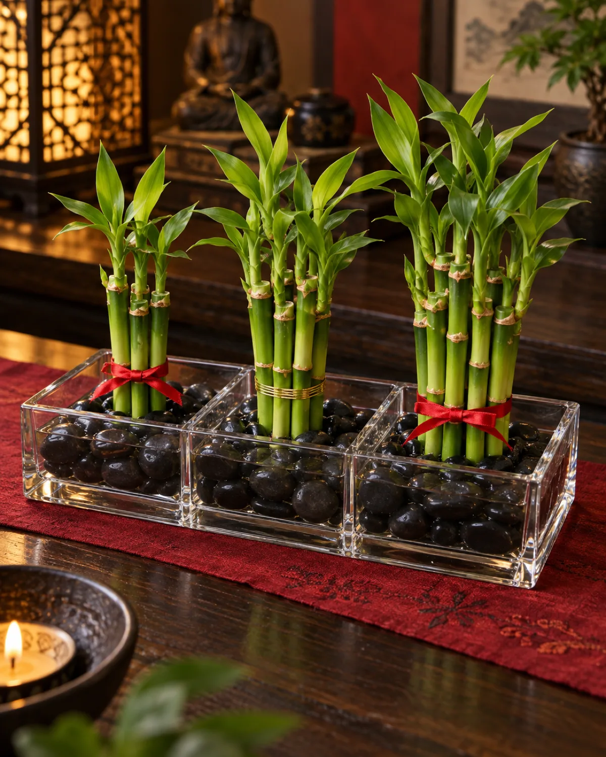 An artistic arrangement of lucky bamboo stalks in different numbers in a clear rectangular glass tray with groups of 3 stalks 5 stalks and 8 stalks each tied with a small red ribbon smooth black river stones at the bottom on a dark wooden table with a subtle red cloth warm ambient lighting