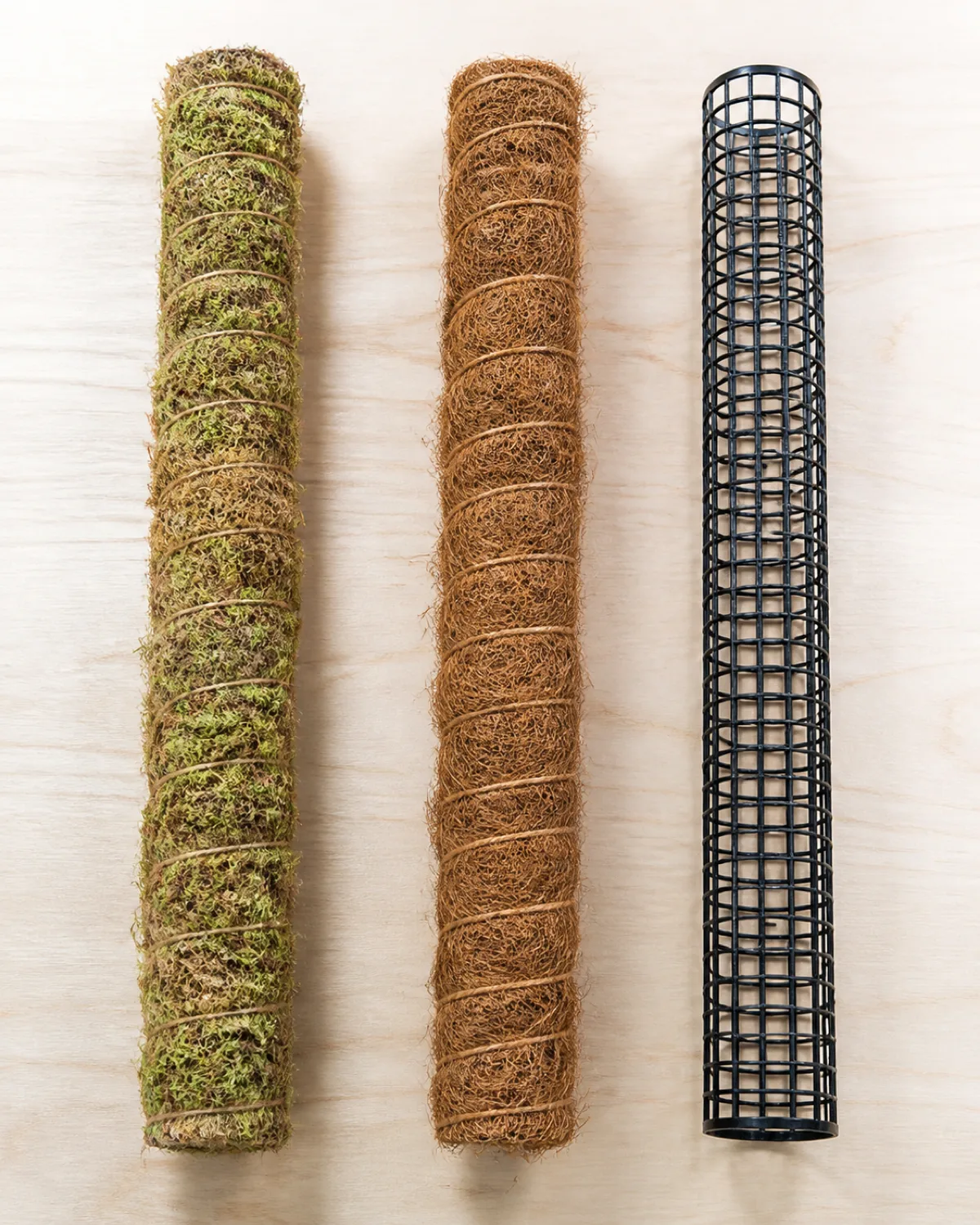 Three types of plant support poles laid side by side on a light wooden table for comparison a sphagnum moss pole wrapped in green-brown fibrous moss left a brown coco coir pole with coarse fiber texture center and a black metal mesh tube with grid pattern right each approximately the same height clean white background product comparison style overhead natural light