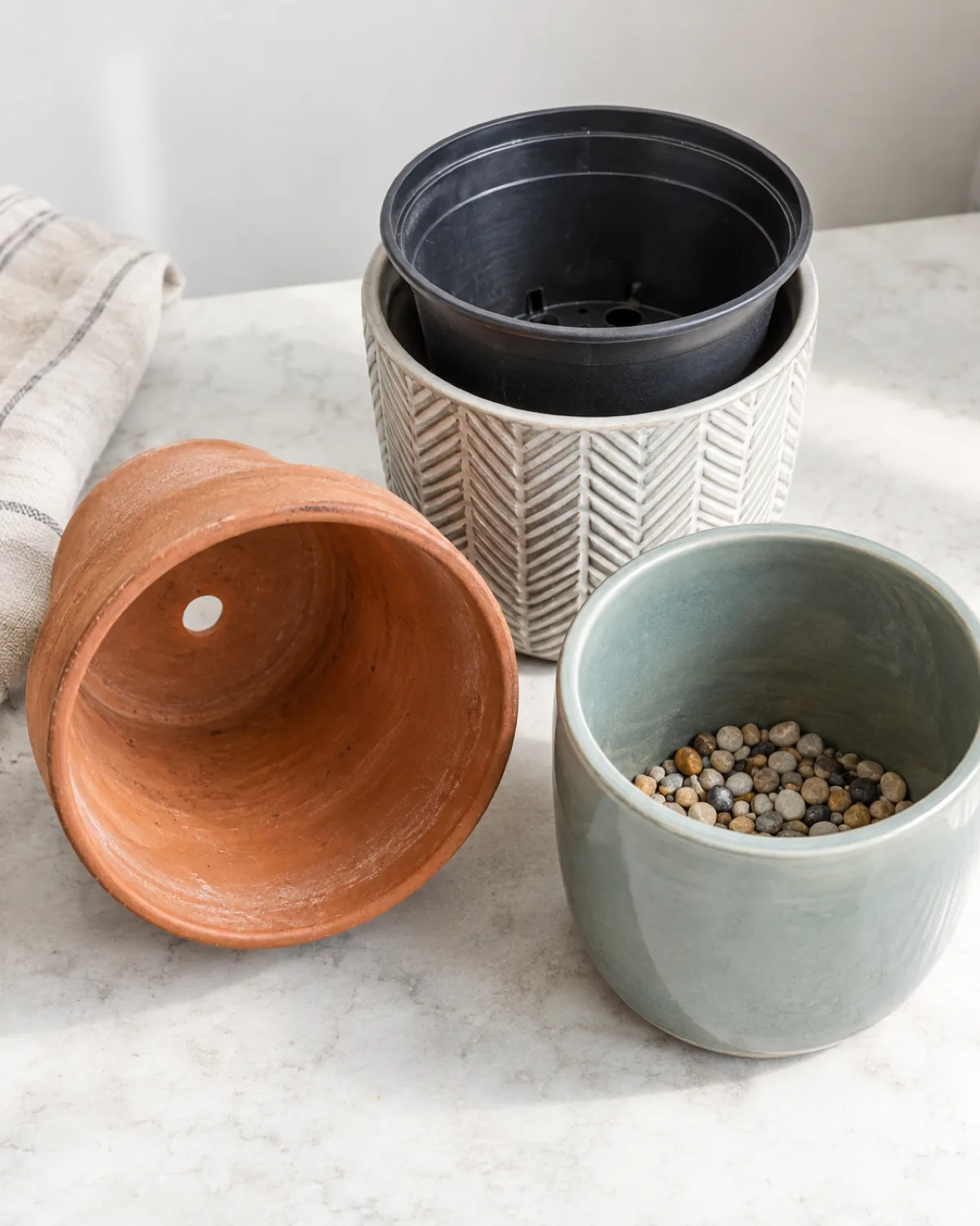 Three plant pots showing drainage comparison a terracotta pot with visible drainage hole a plastic nursery pot inside a decorative cache pot with an air gap and a ceramic pot without drainage with a pebble layer visible inside