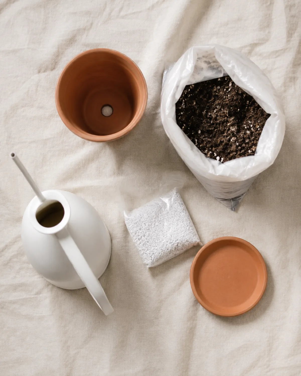 Flat lay on linen surface showing essential beginner plant supplies including a terracotta pot with visible drainage hole a small bag of indoor potting mix a white watering can a bag of perlite and a plain saucer arranged neatly with natural overhead light