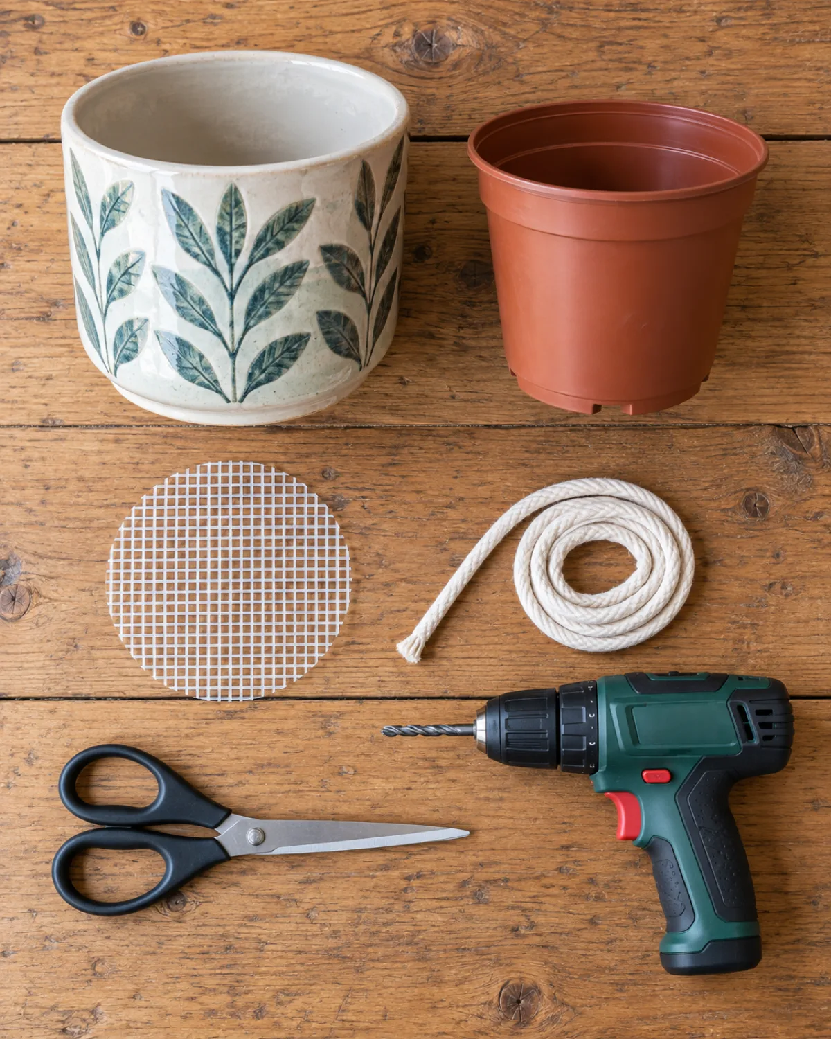 A flatlay arrangement showing materials for a DIY self watering pot conversion on a wooden table including a decorative ceramic cachepot a plastic nursery pot a piece of cut plastic mesh a length of white cotton rope scissors and a small drill all arranged in knolling style
